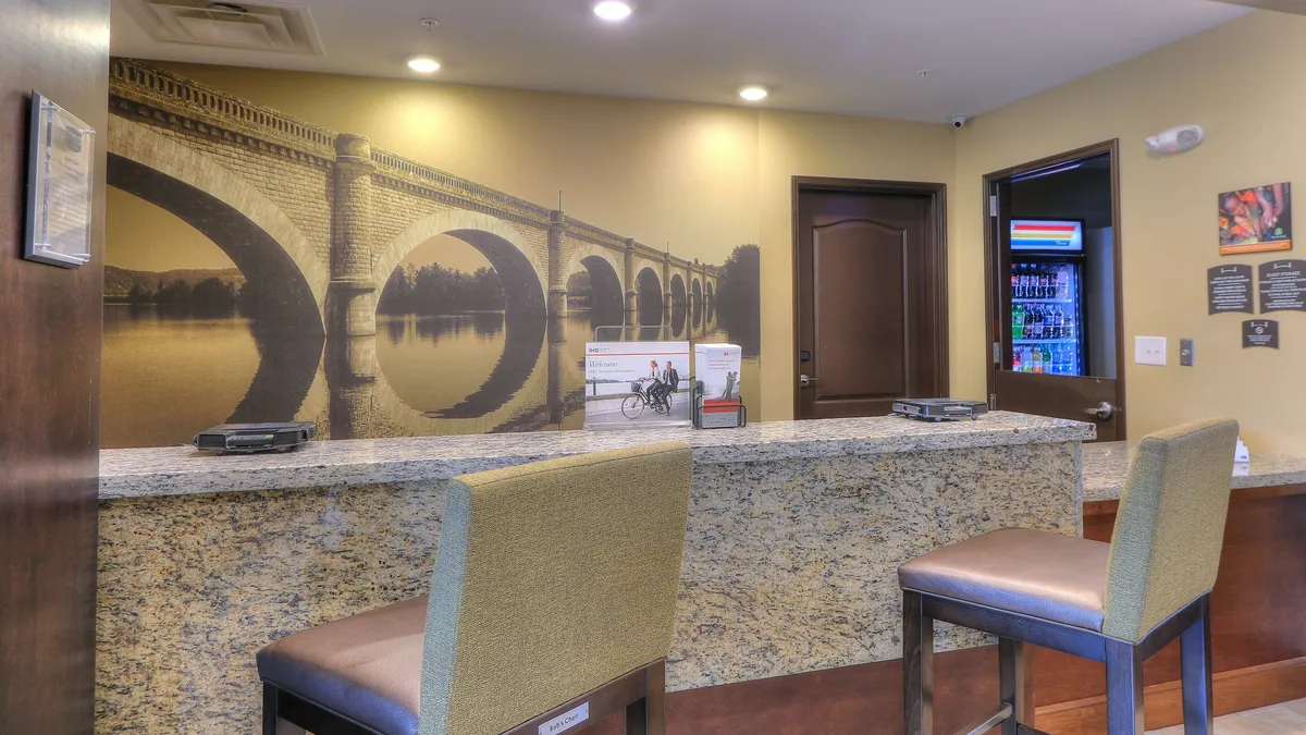 The image shows the hotel's interior reception area, featuring a polished granite counter, a large mural depicting a local bridge, comfortable seating, and a brochure stand with information about local attractions. Staff are seen engaging with guests, providing friendly service and sharing insights about area activities.