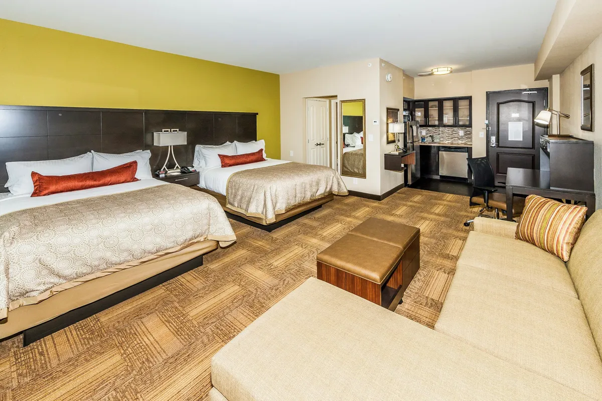 This inviting studio suite features two plush queen beds, a comfortable sofa seating area, a well-lit work desk, and a convenient kitchenette, all arranged in an open, cozy space designed for relaxation and productivity.
