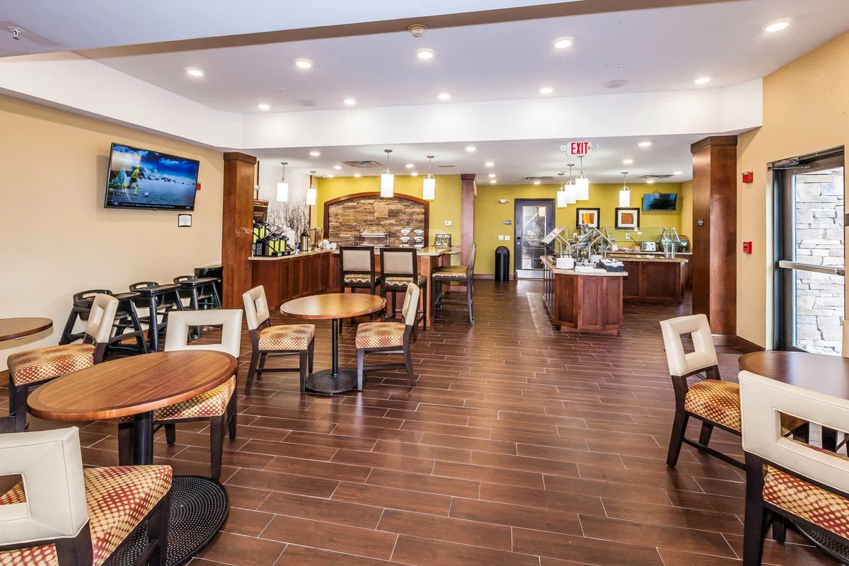 A stylish and relaxed dining area with round tables, high stools at a bar, a decorative stone-accent wall, and a mounted TV. The space is designed for casual dining, social interaction, and watching TV, creating a comfortable environment for meals or evening socials.