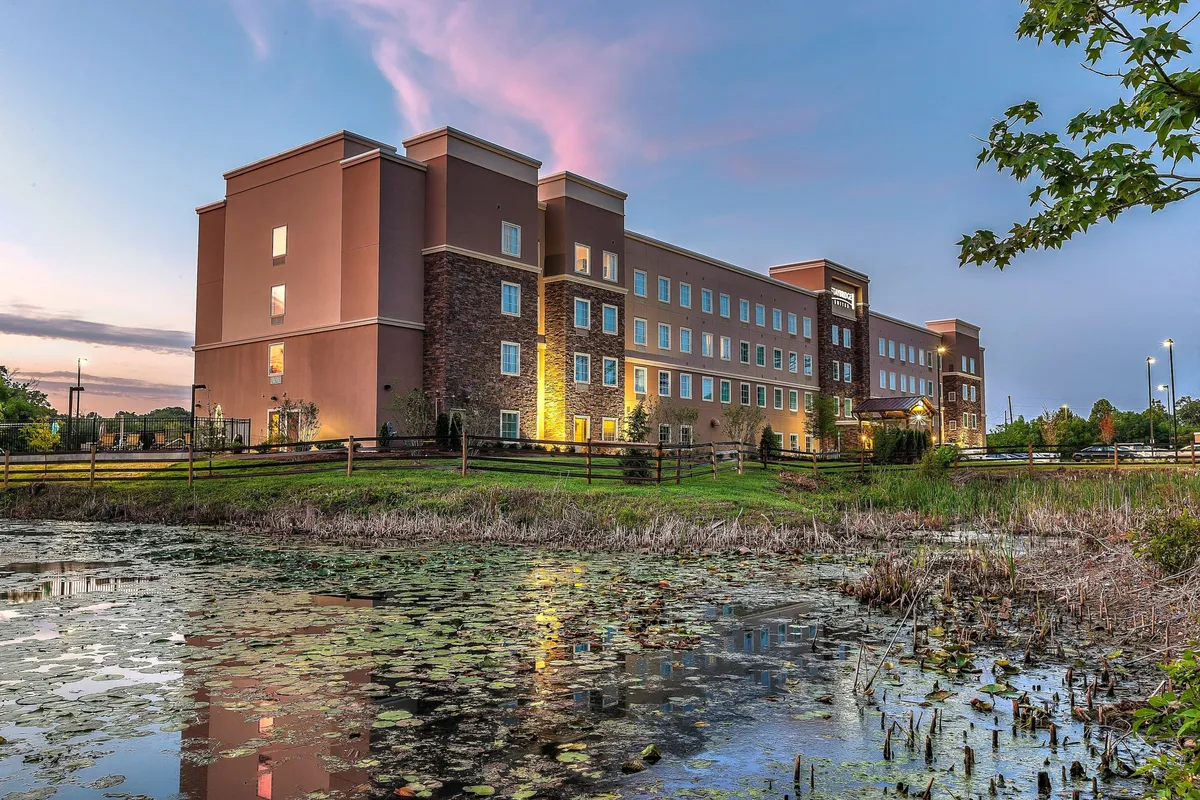 The hotel exterior is seen beside a serene pond, set under a wide, partly cloudy sky. The tranquil setting invites guests to enjoy peaceful moments outdoors, with natural scenery enhancing the sense of relaxation. The view suggests space for evening strolls or unwinding by the water.