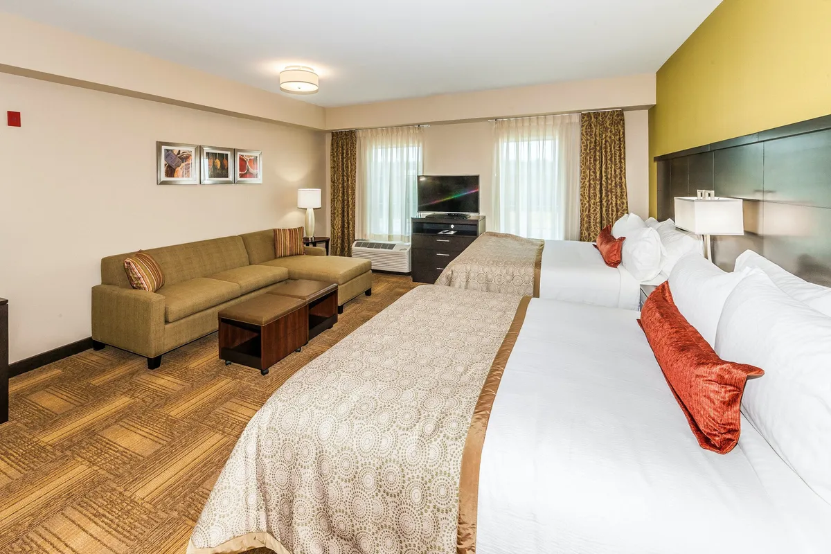 This image showcases a spacious studio suite interior with two queen beds adorned with decorative pillows, a comfortable sofa seating area, a television, and eye-catching artwork on the wall. Large windows allow natural light to fill the space, while blackout curtains are subtly visible, ensuring both brightness and privacy as needed. The atmosphere is homely, inviting, and perfectly suited for relaxation or entertainment.