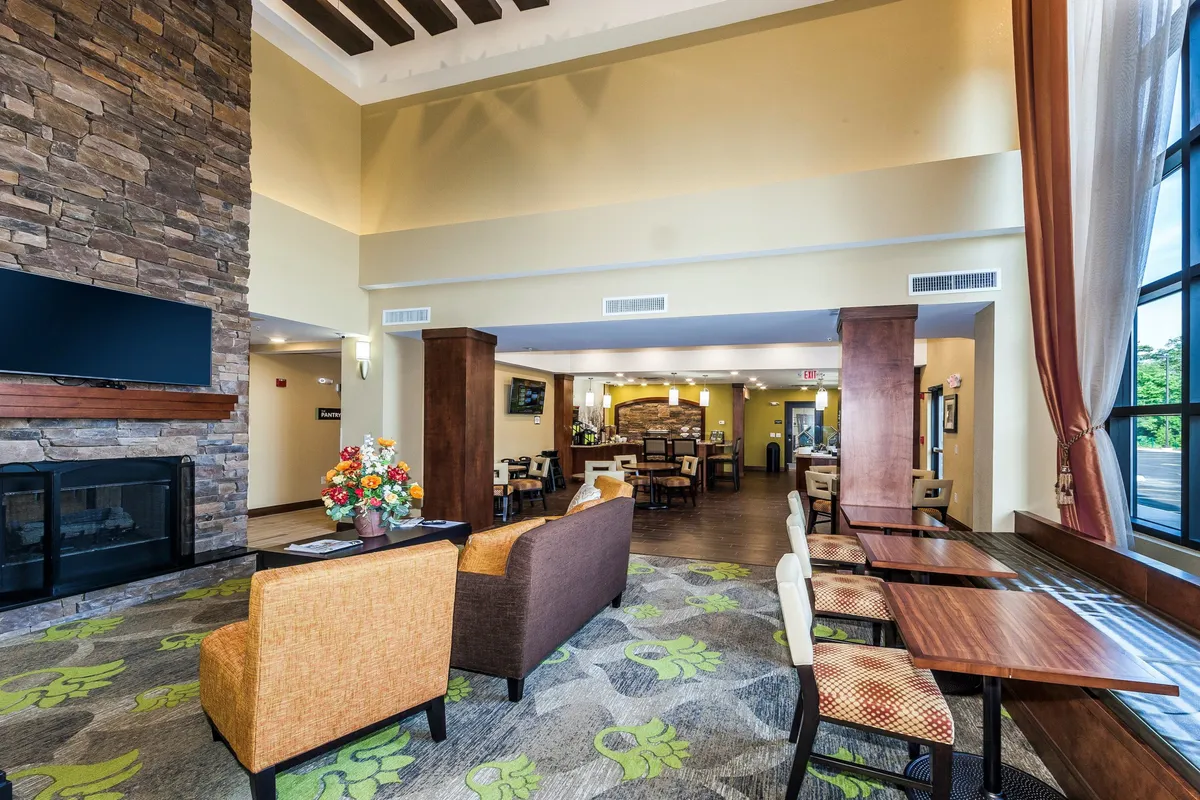 A stylish lounge area with a cozy fireplace, plush seating, decorative flowers, and expansive windows letting in abundant natural light. The space invites relaxation, socialization, or quiet moments for reading or working on a laptop, echoing the homely comfort described in the section.