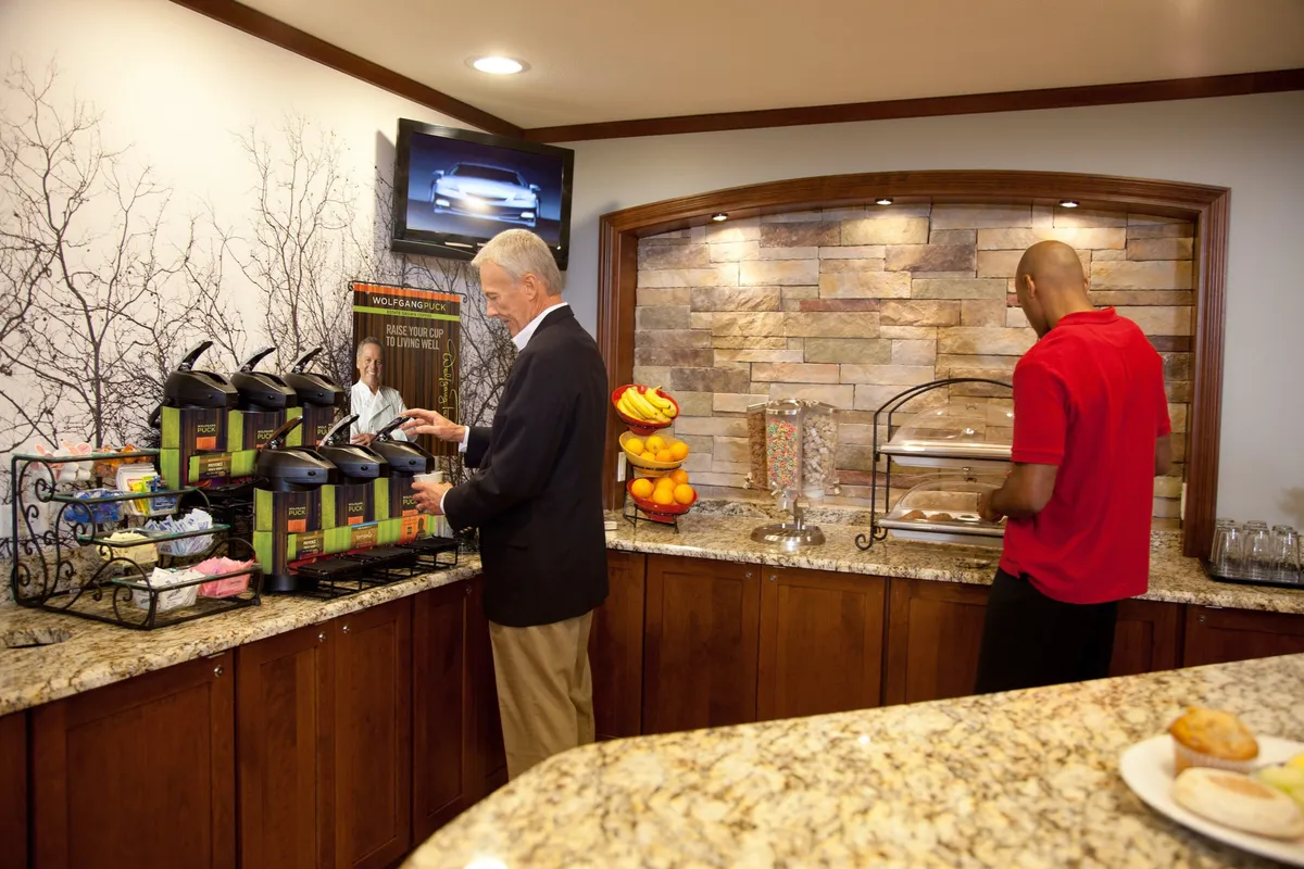 A bright, casual breakfast area featuring a coffee station, fresh fruit display, and assorted breakfast items set up on counters. The space includes comfortable seating and a visible TV screen, inviting guests to relax and enjoy their morning routines.